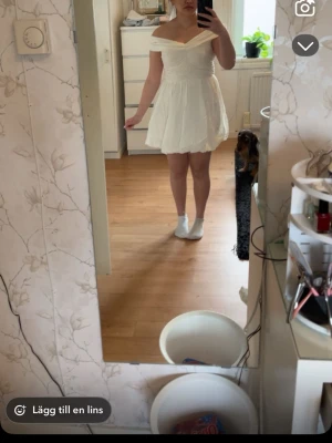 Studentklänning - Söt vit kortklänning med offshoulder-design och lätt puffig kjol. Klänningen har en omlottliknande överdel och markerad midja som ger en feminin siluett. Perfekt för sommarens alla tillfällen.