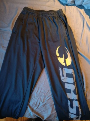 Svarta träningsbyxor från GASP - Säljer ett par svarta Träningsbyxor från GASP med elastisk midja och stort tryck längs benet i vitt och gult. Byxorna är tillverkade i ett ventilerande syntetmaterial som passar perfekt för gymmet eller annan träning.