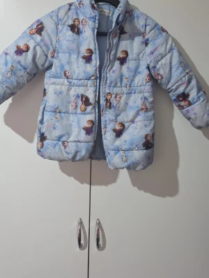 Blå Frozen pufferjacka med motiv - Superfin blå pufferjacka med motiv från Disney Frozen. Jackan har tryck med Elsa, Anna och Olaf över hela ytan och är vadderad för extra värme. Perfekt för kalla vinterdagar och har en mysig känsla. Dragkedja framtill och stora fickor.