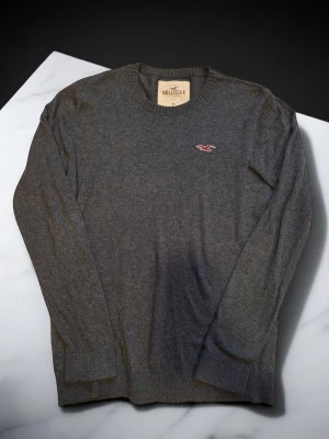 Grå stickad tröja från Hollister - Mörkgrå stickad tröja från Hollister med rund halsringning och ribbade muddar. Tröjan har en liten broderad Hollister-logga i rött och vitt på bröstet. Perfekt för lager-på-lager och enkel att matcha med jeans.