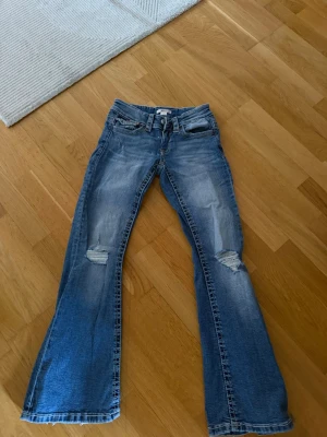 Blå bootcut jeans med slitningar - Säljer ett par blå bootcut jeans med slitningar på båda knäna och snygga detaljer på bakfickorna. Jeansen har en klassisk femficksmodell och är tillverkade i denim med lätt tvättad look. Perfekta för en avslappnad och trendig stil.