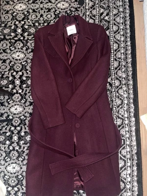 Vinröd ullrock med skärp från Samsoe - Stilren vinröd rock från Samsoe med klassisk krage, knäppning framtill och matchande midjeskärp. Tillverkad i ullblandning med slät innerfoder. Perfekt för dig som vill ha en elegant och tidlös look.