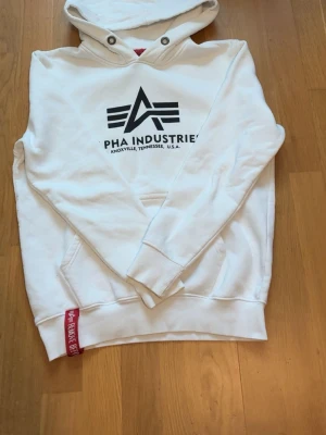 Vit hoodie från Alpha Industries - Snygg vit hoodie från Alpha Industries med svart logga och text på bröstet. Klassisk känguruficka framtill, ribbade muddar och huva med dragsko. Röd Alpha Industries-tag vid nederkanten. Perfekt för en avslappnad och trendig stil.