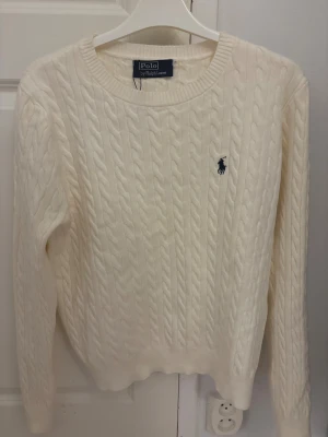 Vit kabelstickad tröja Polo Ralph Lauren - Kabelstickad vit tröja från Polo Ralph Lauren med klassisk blå broderad logga på bröstet. Rund halsringning och ribbade muddar. Mjuk och skön känsla, perfekt för kyliga dagar. Långärmad och i normal passform.