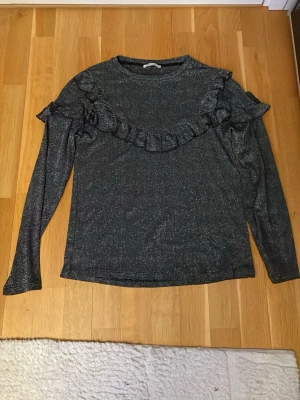 Glittrig svart topp med volang - Snygg svart långärmad topp med glittrigt tyg och volangdetaljer över bröstet och axlarna. Rund halsringning och figurnära passform. Perfekt för dig som vill sticka ut lite extra. Materialet känns stretchigt och bekvämt.