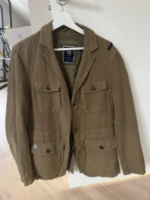 Olivgrön kavaj från Hansen Jacob  - Snygg olivgrön kavaj från Henry Choice med fyra framfickor och klassisk krage. Kavajen har dekorativa knappar och patchdetalj på insidan. Tillverkad i ett mjukt bomullstyg som ger en avslappnad look. Pris 1499kr, passar M/L