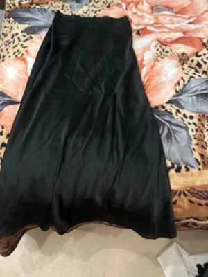 Svart långkjol i satin - Stilren svart långkjol i glansigt satinmaterial. Kjolen har en mjuk och följsam passform som ger en snygg siluett. Perfekt för dig som gillar minimalistisk och elegant stil. Behöver strykas. 