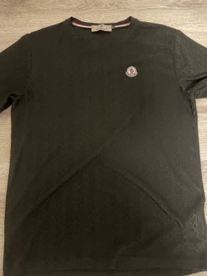 Svart Moncler t-shirt med diskret mönster - Den är XL i barn storlek så kanske S-M i vuxen strorlek. Säljer en svart Moncler t-shirt i storlek XL med diskret geometriskt mönster i tyget och Moncler-logga på bröstet. Klassisk rund halsringning och korta ärmar. Perfekt för dig som gillar stilrena plagg med exklusiv känsla. 