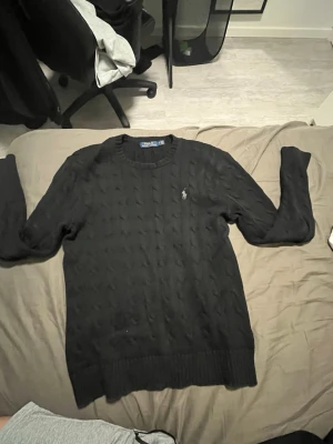 Svart kabelstickad tröja Polo Ralph Lauren - Kabelstickad svart tröja från Polo Ralph Lauren med rund halsringning och diskret broderad logga på bröstet. Tröjan har långa ärmar och ribbade muddar vid ärmslut och nederkant. Perfekt för en stilren och klassisk look.