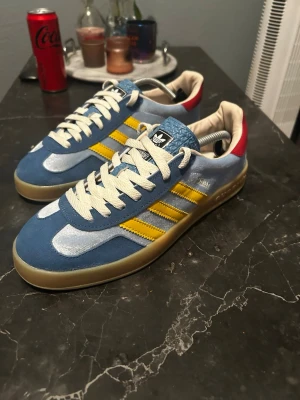 Adidas blå och gula sneakers - Snygga Adidas x gucci sneakers i blå mocka och textil med gula ränder på sidorna och röda detaljer vid hälen. Skorna har vita snören, gummisula och klassisk Adidas-logga på plösen. Perfekta för dig som gillar retrostil och vill sticka ut med färg.