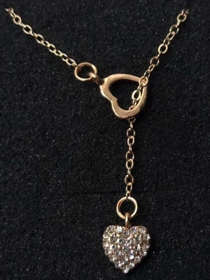 Guldigt halsband med hjärthängen - Snyggt halsband i guldfärgad metall med två hjärtformade hängen, varav det ena är dekorerat med glittrande stenar som ger en pärl-liknande effekt. Kedjan är tunn och elegant, perfekt för att lyfta vilken outfit som helst. 