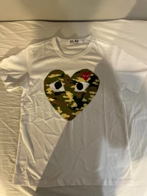 Vit Comme des Garçons PLAY t-shirt - Vit t-shirt från Comme des Garçons PLAY med ett stort hjärta i camouflage-mönster och ögon på bröstet samt ett litet rött hjärta ovanför. Klassisk rund hals och korta ärmar. Perfekt statement-plagg för dig som gillar streetwear.