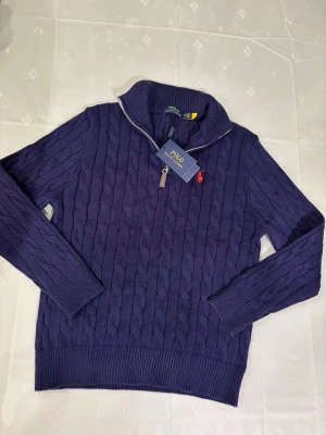 Mörkblå kabelstickad tröja Polo Ralph Lauren - Mörkblå kabelstickad tröja från Polo Ralph Lauren med halv dragkedja och hög krage. Tröjan har det klassiska röda Polo-logot broderat på bröstet och ribbade muddar vid ärmslut och nederkant. Perfekt för lager på lager och snygg till jeans.