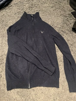 Mörkblå ziptröja från GANT - Mörkblå ziptröja från GANT i extra fin lammull. Tröjan har hög krage, dragkedja framtill och diskret GANT-logga broderad på bröstet. Perfekt för lager på lager och ger en clean look till din outfit.