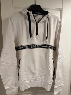 Vit hoodie från Emporio Armani EA7 - Snygg vit hoodie från Emporio Armani EA7 med svart dragkedja och svarta snören i huvan. Dekorativt band med logga tvärs över bröstet och ryggen. Två fickor med dragkedja framtill och ribbade muddar. 