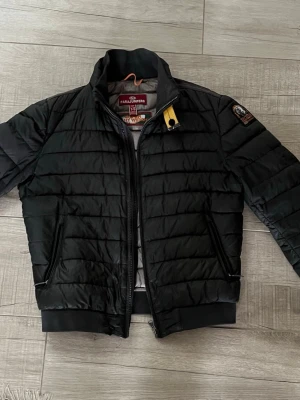 Svart pufferjacka från Parajumpers - Snygg svart Parajumpers superlight-weight med gul detalj vid kragen och patch på ärmen. Jackan har två dragkedjeförsedda fickor, ribbade muddar och quiltad design. Perfekt för kyligare dagar och riktigt stilren look. Bra för våren. Grisch. Mycket bra skick
