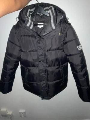 Svart pufferjacka från  Burberry - Snygg svart pufferjacka från Burberry med huva och dragkedja. Jackan har ett randigt innerfoder i grått och vitt samt tryck på ärmen. Perfekt för kalla vinterdagar och har en modern, boxig passform.