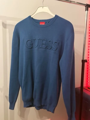 Blå stickad tröja från Guess - Säljer en blå stickad tröja från Guess med broderad logga framtill. Tröjan har rund halsringning, långa ärmar och ribbade muddar vid ärmslut och nederkant. Perfekt för dig som gillar stilrena och klassiska plagg.
