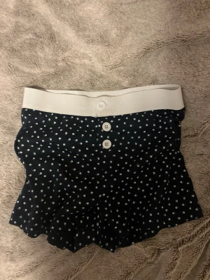 Mörkblå pyjamasshorts med blommor - Söta mörkblå pyjamasshorts med små vita blommor och bred vit midjeresår. Shortsen har dekorativa knappar framtill och volangkant nedtill för en extra gullig look. Perfekta för mysiga kvällar hemma. 