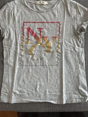 Grå NYC t-shirt från H&M - Säljer en grå t-shirt från H&M med ett stort NYC-tryck i rött och gult framtill. T-shirten har rund halsringning och korta ärmar. Tillverkad i mjuk bomull, perfekt för en avslappnad och cool stil.
