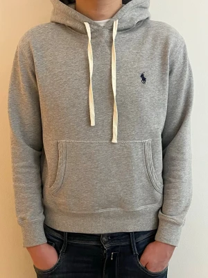 Grå hoodie från Polo Ralph Lauren - Klassisk grå hoodie från Polo Ralph Lauren med vit dragsko i huvan och mörkblå logga broderad på bröstet. Hoodien har känguruficka framtill, ribbade muddar och är tillverkad i mjuk bomullsblandning. Perfekt för en avslappnad och stilren look.