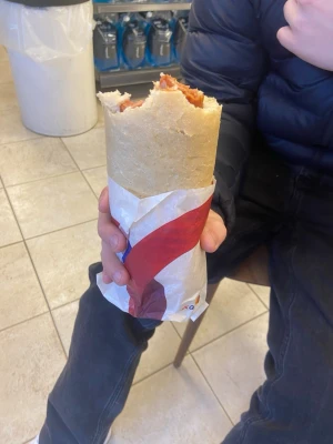 Stor rulle kebab i tunnbröd - En rejäl kebabrulle inslagen i papper, fylld med kebabkött och sås. Tunnbrödet är ljust och mjukt, och rullen är stor nog att hålla med båda händerna. Perfekt för dig som gillar matiga snacks eller snabbmat.