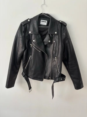 Svart biker skinnjacka  - Snygg svart biker skinnjacka från Noysi May med silvriga dragkedjor, axelklaffar och bälte i midjan. Klassisk modell med asymmetrisk dragkedja och flera fickor framtill. 
