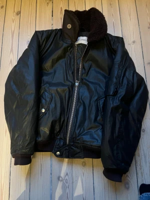 Svart bomberjacka från Our Legacy - Snygg svart bomberjacka från Our Legacy med glansig finish, dragkedja framtill och två snedställda fickor med knapp. Jackan har ribbade muddar vid ärmar och midja samt en hög krage med mjukt foder. Perfekt för dig som vill ha en stilren och trendig look.