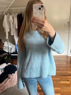 Ljusblå stickad tröja - Mysig ljusblå stickad tröja med avslappnad passform och rund hals. Tröjan har breda ribbade muddar vid ärmslut och nederkant, vilket ger en trendig look. Perfekt att matcha med jeans för en chill vardagsstil.