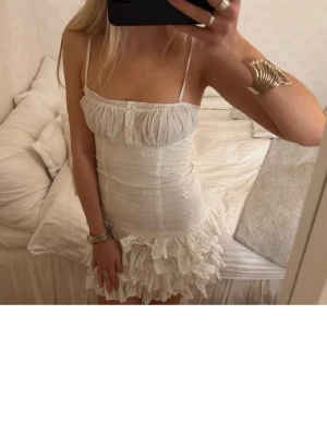 Vit kort volangklänning med spets - Supersöt vit kort klänning med tunna axelband, knappar framtill och volangdetaljer längst ner. Klänningen har broderade detaljer och ärmlös design som ger en romantisk och somrig känsla. Perfekt för dig som vill ha en feminin och trendig look. Perfekt till studenten!