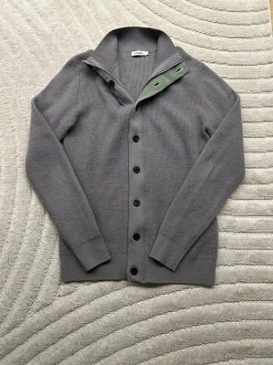 J.Lindeberg stickad cardigan - stl S - Snygg grå stickad kofta från Aspesi med svarta knappar och ribbade muddar. Koftan har en klassisk krage och en unik detalj med ljusgrönt tyg på insidan av knappslån. Perfekt för lager på lager och stilrena outfits.