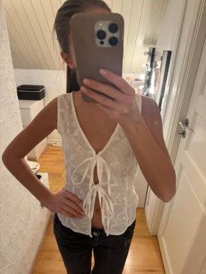 Vit blommig spetslinnetopp med knyt - Säljer en söt vit ärmlös linnetopp med blommigt spetsmönster och två knyt framtill. Toppen har v-ringning och är lätt transparent, perfekt för sommarens alla outfits. Superfin detalj med de broderade blommorna och den luftiga känslan.