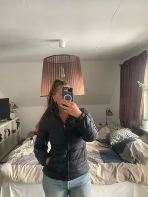 Svart pufferjacka med huva - Snygg svart pufferjacka med dragkedja och huva. Jackan har en quiltad design och två fickor framtill. 