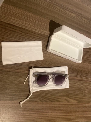 Transparenta solglasögon från Chimi - Snygga solglasögon från Chimi med transparent båge och mörklila glas. Rektangulär form som ger en modern vibe. Kommer med vit förvaringspåse och hårt etui. Perfekta för dig som vill sticka ut med stilrena accessoarer. Glasögonen har små defekter men inget som syns vid användning.