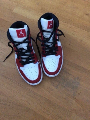 Air Jordan 1 High röd/vit/svart - Säljer ett par Air Jordan 1 High sneakers i klassisk röd, vit och svart färgkombination. Skorna har svart swoosh, svarta skosnören och perforerad tåbox. Tillverkade i läder med högt skaft och ikoniska Air Jordan-detaljer på plös och sida.