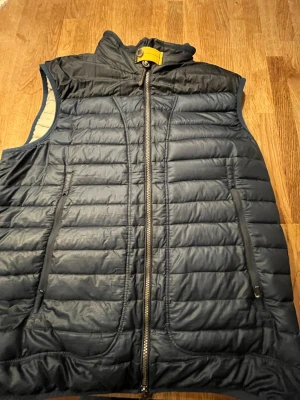 Mörkblå dunväst från Parajumper - Snygg mörkblå dunväst från Parajumper med gul detalj vid kragen och två dragkedjeförsedda fickor. Västen har quiltad design och hög krage för extra värme. Perfekt för lager-på-lager och outdoor-vibe. Där är ett litet hål på ryggen och ett litet hål vi den ena fickan! Stl XL men passar stl M och L