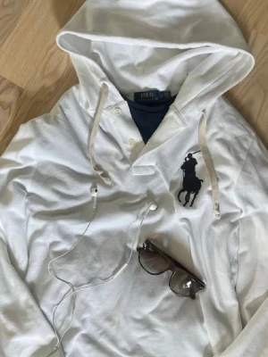 Vit big pony hoodie från Ralph Lauren.  - Vit hoodie från Polo Ralph Lauren med stor svart logga på bröstet. Tröjan har huva med snörning, knappar vid halsen och är tillverkad i bomull. Långärmad och klassisk passform, perfekt för en clean och stilren look. Storlek S i bra skick utan defekter. 