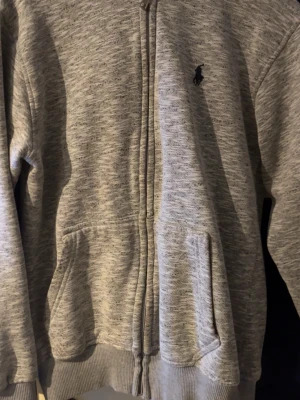 Gråmelerad hoodie från Polo Ralph Lauren - Klassisk gråmelerad hoodie från Polo Ralph Lauren med dragkedja framtill och två fickor. Broderad logga på bröstet och mjukt material i bomullsmix. Perfekt för en chill och avslappnad stil.