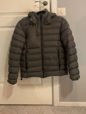 Mörkgrå dunjacka med huva från Colmar - Snygg mörkgrå dunjacka från Save The Duck med avtagbar huva och två dragkedjeförsedda fickor. Jackan har quiltad design och rund Save The Duck-logga på ärmen. Perfekt för kalla vinterdagar och riktigt skön att bära.