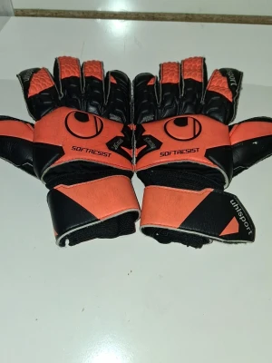 Uhlsport målvaktshandskar orange/svart - Säljer ett par målvaktshandskar från Uhlsport i orange och svart med breda kardborreband runt handleden. Handskarna har greppvänlig insida och är märkta med 'SOFTRESIST'. Perfekta för fotbollsträning eller match.