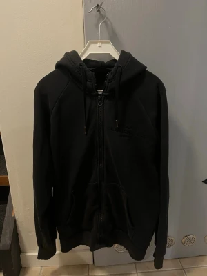 Svart hoodie från Peak Performance - Säljer en svart hoodie från Peak Performance med dragkedja framtill och justerbar huva med snören. Hoodien har fickor på sidorna och diskret logga på bröstet. Tillverkad i mjukt och bekvämt material, perfekt för en avslappnad stil. Lite missfärgad runt dragkedjan och lite sliten i sömändarna