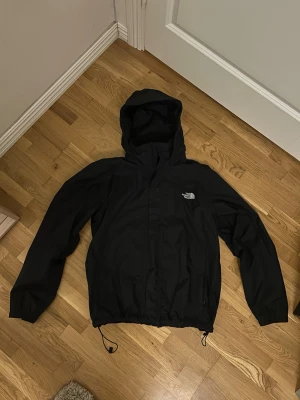 Svart vindjacka från The North Face - Svart vindjacka från The North Face med huva och dragkedja framtill. Jackan har meshfoder på insidan, justerbar nederkant med dragsko och två sidofickor. Perfekt för blåsiga dagar och enkel att matcha med olika outfits.