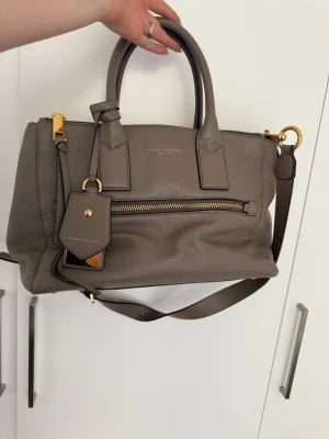 Beige/grå handväska från Marc Jacobs - Snygg grå handväska från Marc Jacobs i mjukt läder med guldfärgade metalldetaljer. Väskan har både handtag och avtagbar axelrem, samt en praktisk ytterficka med dragkedja och en dekorativ tag. Perfekt för dig som gillar stilrena accessoarer.