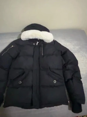 Svart dunjacka från Moose Knuckles - Säljer en svart dunjacka från Moose Knuckles med vit fuskpäls på huvan och ikonisk metallogo på ärmen. Jackan har dragkedja, flera fickor med dragkedja och tryckknappar samt en rejäl, vadderad design som håller dig varm under kalla dagar.