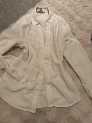 Vit skjorta med knappar - Säljer en stilren vit skjorta med klassisk krage och knäppning framtill. Skjortan har lång ärm och är tillverkad i ett lätt och mjukt material som ger en snygg passform. Perfekt att styla till olika outfits.
