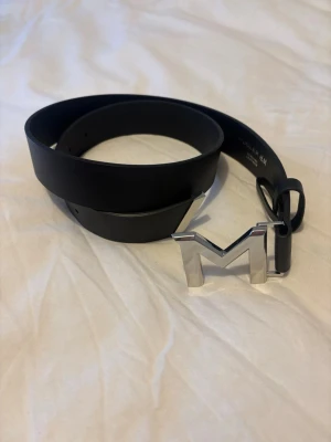 Svart läderbälte Mugler H&M - Snyggt svart bälte från Mugler H&M i äkta läder med en stor, silverfärgad M-formad metallspänne. Perfekt accessoar för att lyfta din outfit med en cool detalj. Bältet har en stilren och modern design.