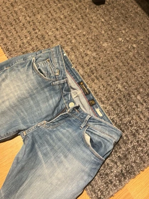 Ljusblå raka jeans från Replay - Säljer ett par ljusblå jeans från Replay med klassisk femficksmodell och raka ben. Jeansen har snygga slitningar och kontrastsömmar samt knappgylf. Perfekta för en avslappnad och trendig look.