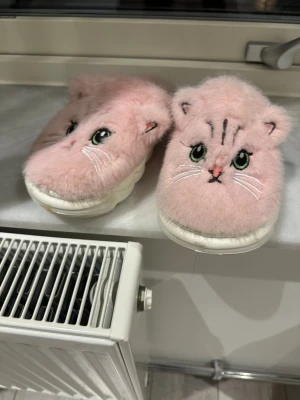 Rosa fluffiga tofflor med kattmotiv - Supersöta rosa tofflor med mjuk, fluffig yta och broderat kattansikte framtill. Tofflorna har vita sulor och små öron som sticker upp, vilket ger en gullig look. Perfekta för att hålla fötterna varma hemma under kalla dagar. Storlek 36-37