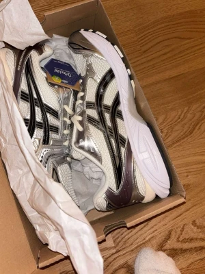 Asics silvervita sneakers - Snygga sneakers från Asics i silver och vitt med svarta detaljer. Skorna har en sportig design med meshpaneler för bra ventilation och Ortholite-innersula för extra komfort. Klassisk snörning och slitstark yttersula. Perfekta för dig som gillar streetwear och sportig stil. Använda 2 gånger 