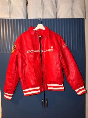 Röd Porsche  skinnjacka - Riktigt snygg röd skinnjacka från Porsche. Jackan har broderade loggor fram och bak, vita detaljer på muddar och krage samt coola patches på ärmarna. Perfekt statement-plagg för dig som gillar streetwear och bilar. OBS DETTA ÄR INTE EN ALD JACKA!
